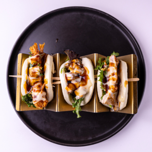 Three bao buns with fried fillings and drizzles of sauce on a black plate.