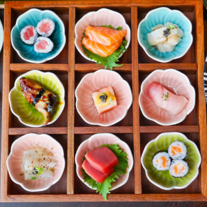 Top-down view of nine small flower-shaped dishes of sushi in a wooden tray.
