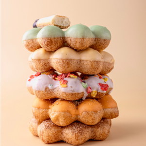 Stack of colorful mochi donuts with various glazes and toppings on a tan background.